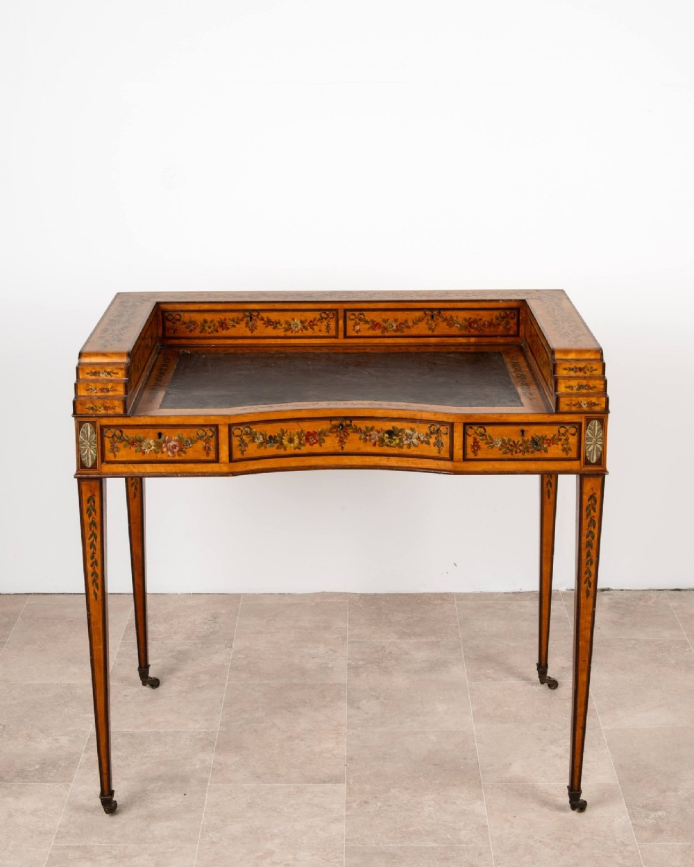 A warm gold satinwood Carlton House writing desk featuring hand-painted floral swags, a dark leather writing surface, and tapered legs on brass castors. View IV