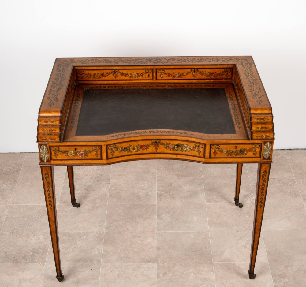 A warm gold satinwood Carlton House writing desk featuring hand-painted floral swags, a dark leather writing surface, and tapered legs on brass castors. View V