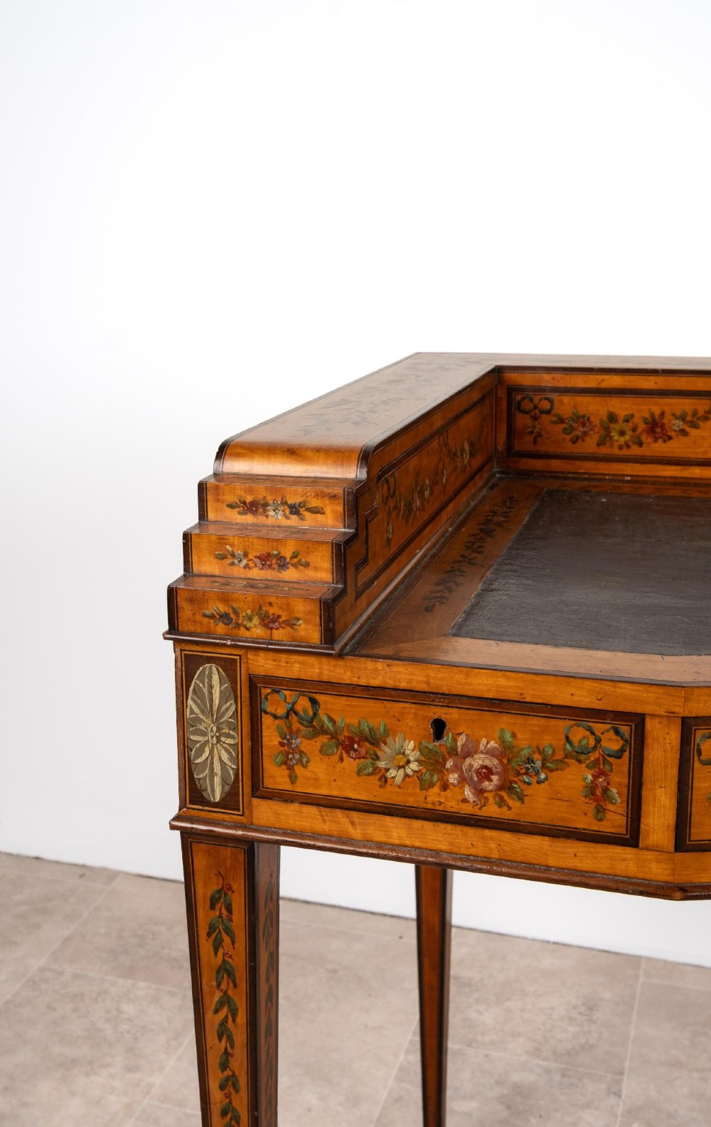 A warm gold satinwood Carlton House writing desk featuring hand-painted floral swags, a dark leather writing surface, and tapered legs on brass castors. View VI