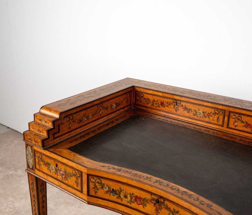 A warm gold satinwood Carlton House writing desk featuring hand-painted floral swags, a dark leather writing surface, and tapered legs on brass castors. View VII