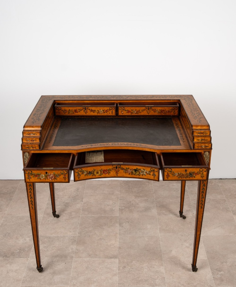 A warm gold satinwood Carlton House writing desk featuring hand-painted floral swags, a dark leather writing surface, and tapered legs on brass castors. View VIII