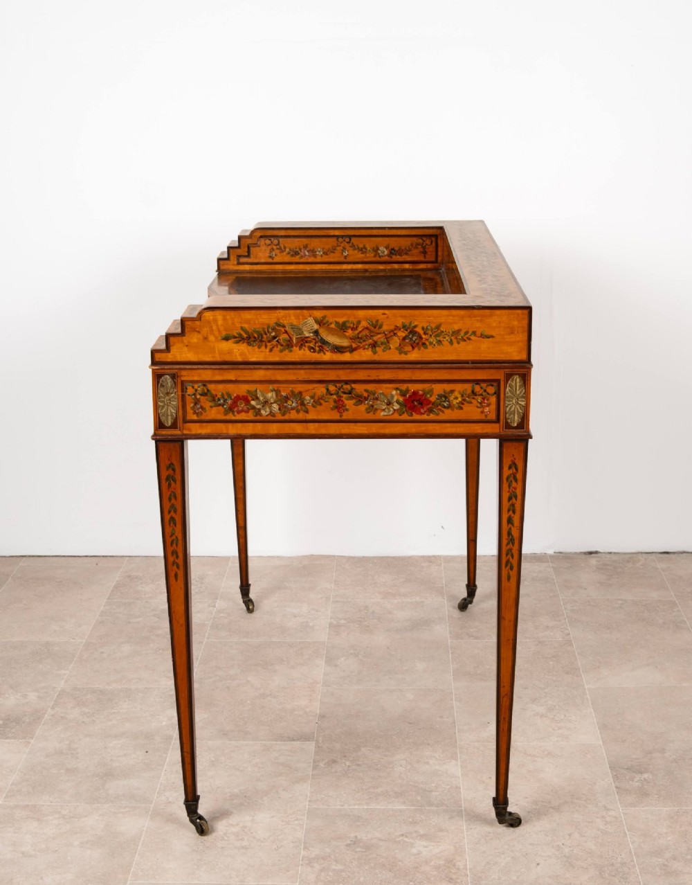 A warm gold satinwood Carlton House writing desk featuring hand-painted floral swags, a dark leather writing surface, and tapered legs on brass castors. View IX