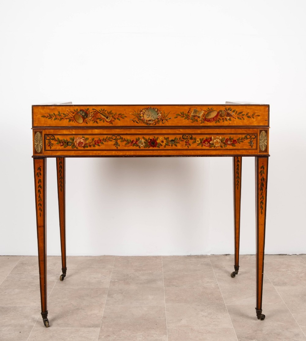 A warm gold satinwood Carlton House writing desk featuring hand-painted floral swags, a dark leather writing surface, and tapered legs on brass castors. View X