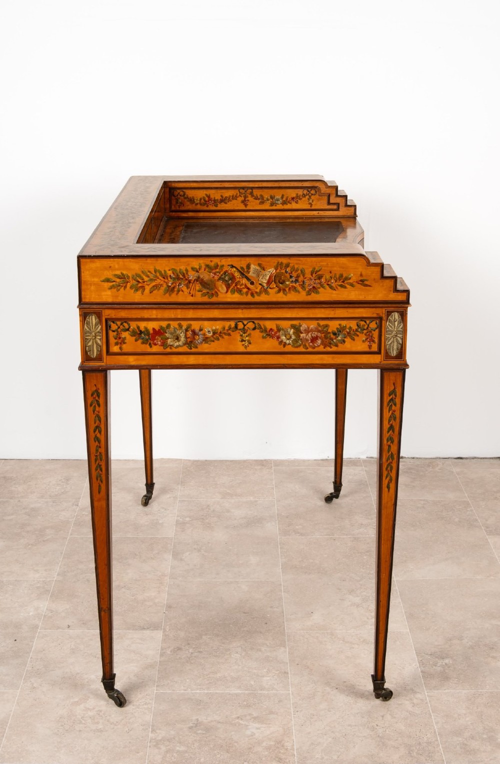 A warm gold satinwood Carlton House writing desk featuring hand-painted floral swags, a dark leather writing surface, and tapered legs on brass castors. View XI