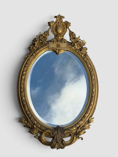 fig 1. An oval Victorian gilt gesso wall mirror featuring an ornate floral crest, decorative scrollwork, and bevelled mirror glass.