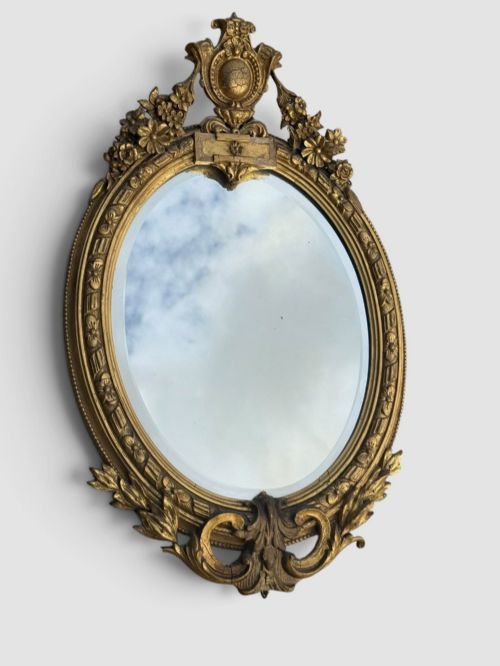 fig 2. An oval Victorian gilt gesso wall mirror featuring an ornate floral crest, decorative scrollwork, and bevelled mirror glass.