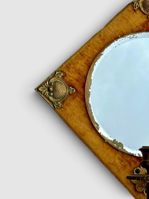 fig 1. A diamond-shaped wall mirror featuring a mustard yellow velvet frame with decorative brass corner mounts, a central round bevelled glass mirror, and an integrated brass candle sconce at the lower vertex.