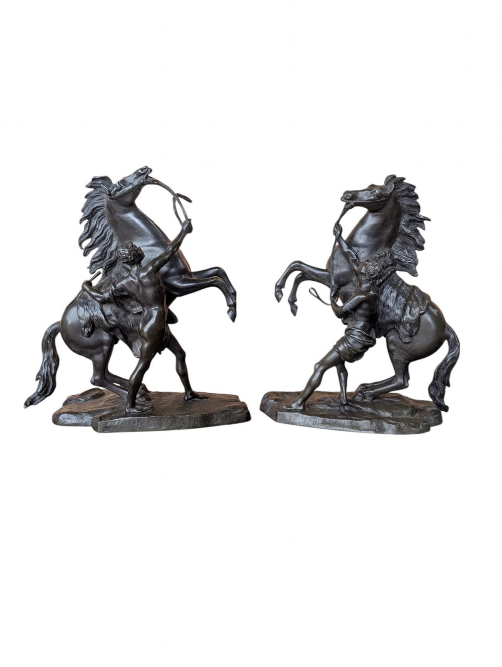 A pair of dark bronze sculptures depicting rearing horses restrained by standing grooms on naturalistic rock bases.