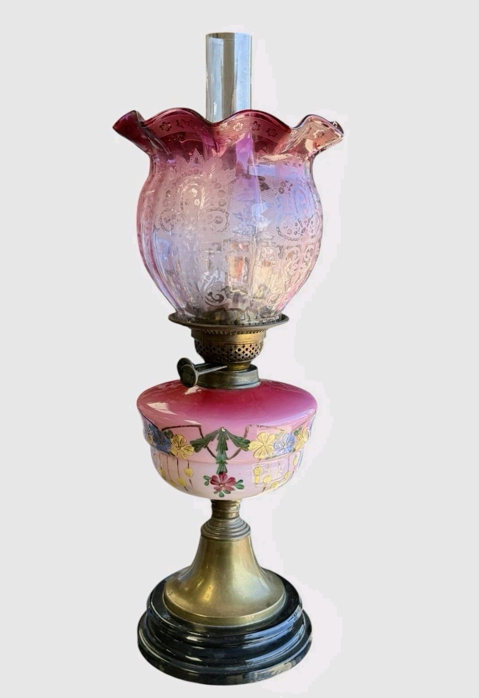 A Victorian cranberry glass oil lamp with a ruffled etched shade, hand-painted floral reservoir, brass burner, and stepped black base.