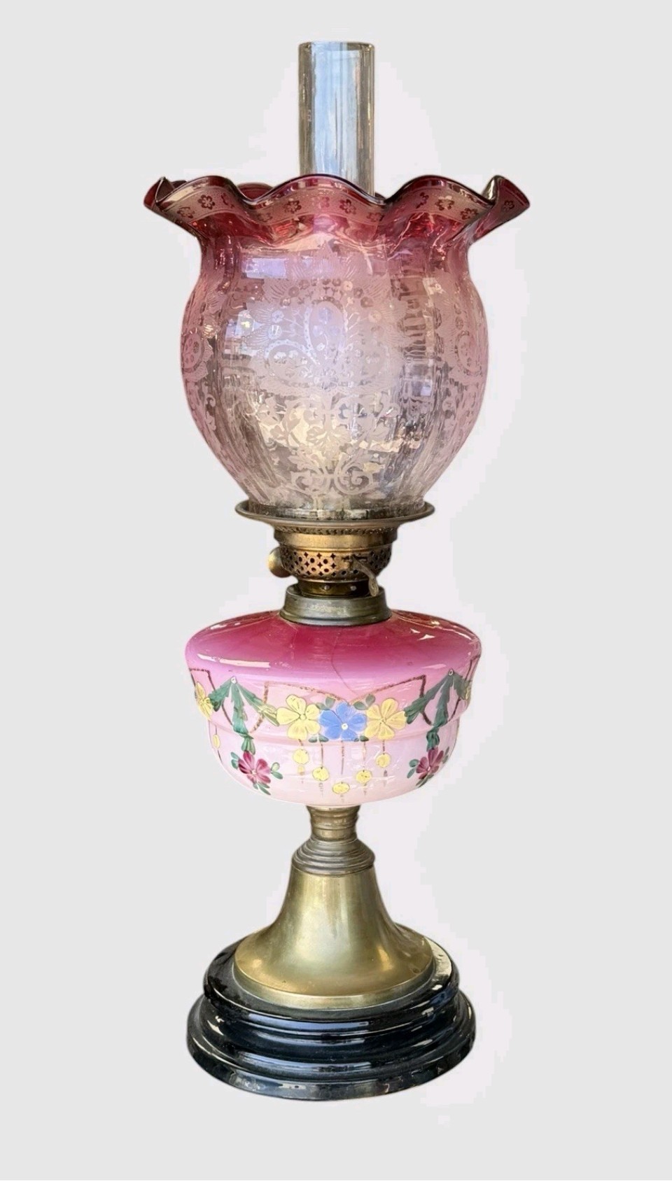A Victorian cranberry glass oil lamp with a ruffled etched shade, hand-painted floral reservoir, brass burner, and stepped black base. View VI