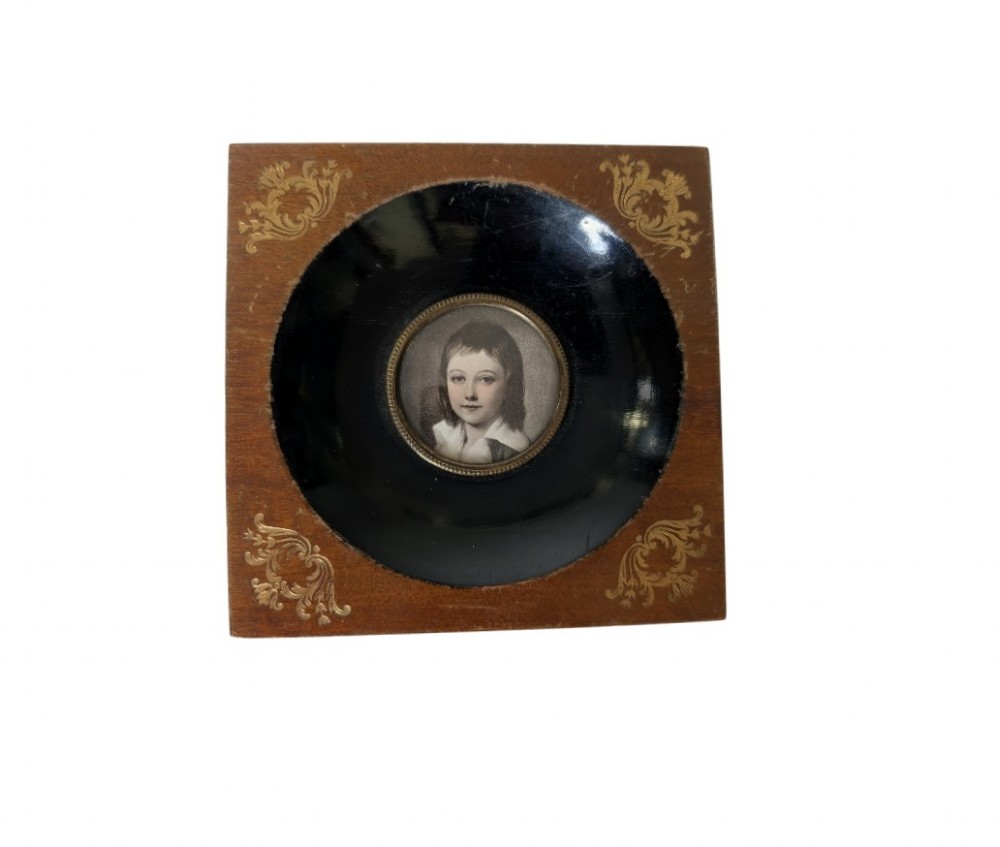 A square wooden frame featuring a dark ebonised circular surround with gilt corner decorations and an inset portrait of a child.