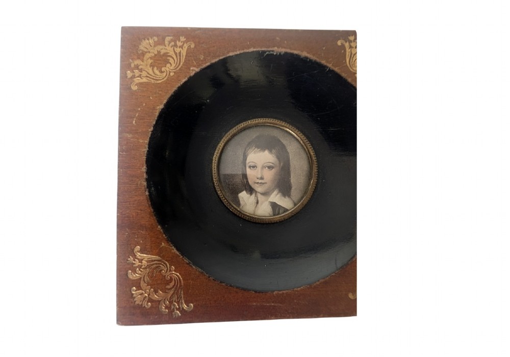A square wooden frame featuring a dark ebonised circular surround with gilt corner decorations and an inset portrait of a child. View III