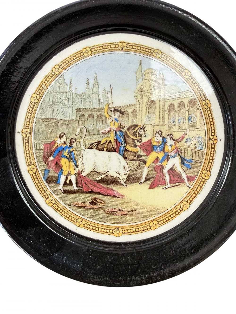 A circular ceramic plate depicting a transfer-printed bullfighting scene, framed within a wide circular black ebonised surround. View IV