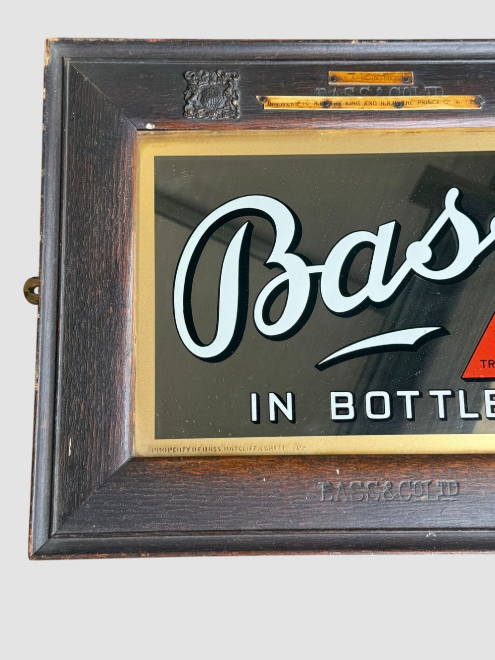 An antique dark oak framed advertising mirror featuring white script reading Bass in Bottle alongside a red triangle logo and brass Royal Warrant badges. View II