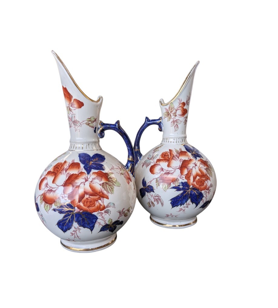 A pair of antique Japanese Imari porcelain pitchers featuring rounded bodies, tall swept spouts, dark blue handles, and hand-painted rust red flowers with gilded accents.