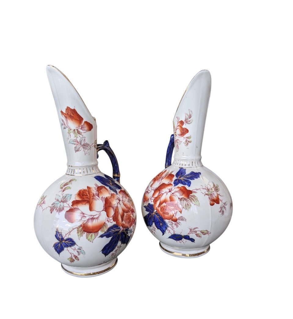 A pair of antique Japanese Imari porcelain pitchers featuring rounded bodies, tall swept spouts, dark blue handles, and hand-painted rust red flowers with gilded accents. View II