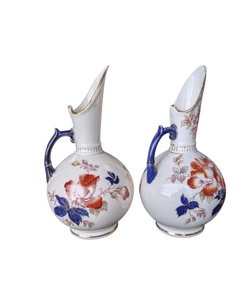 A pair of antique Japanese Imari porcelain pitchers featuring rounded bodies, tall swept spouts, dark blue handles, and hand-painted rust red flowers with gilded accents. View IX
