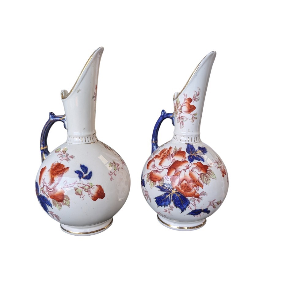 A pair of antique Japanese Imari porcelain pitchers featuring rounded bodies, tall swept spouts, dark blue handles, and hand-painted rust red flowers with gilded accents. View X