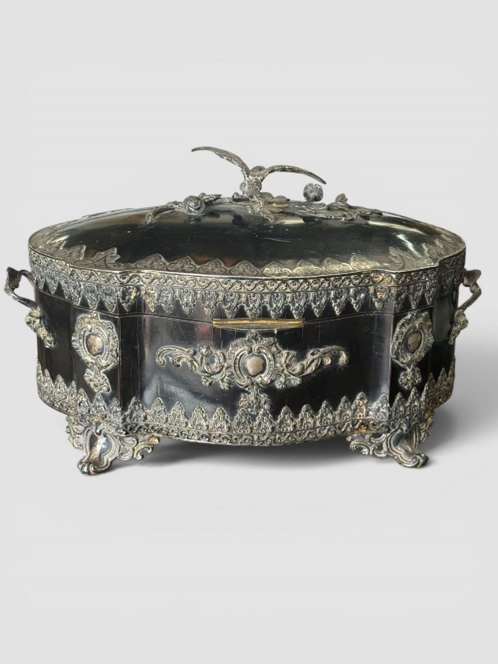 A 19th-century French ebonised serpentine table casket featuring elaborate silvered metal scrollwork mounts, raised feet, and a lid top decorated with a winged bird finial.