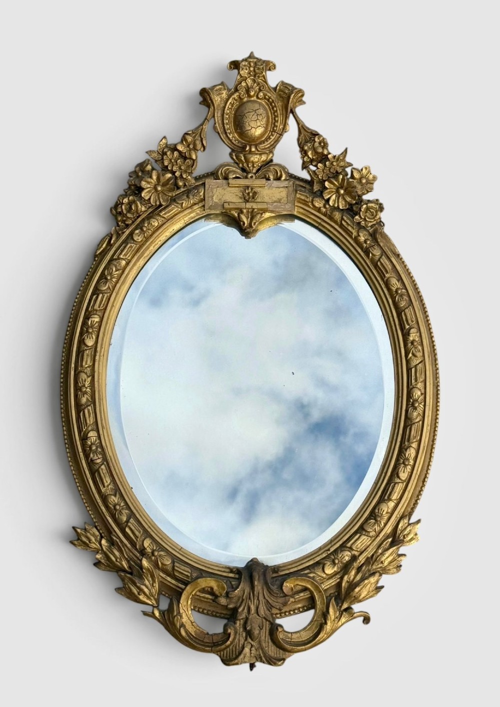 An oval Victorian gilt gesso wall mirror featuring an ornate floral crest, decorative scrollwork, and bevelled mirror glass.