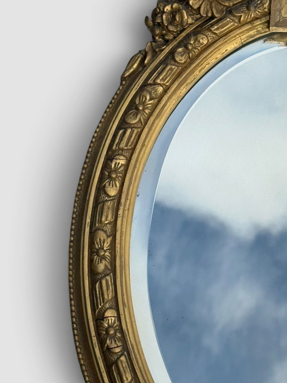 An oval Victorian gilt gesso wall mirror featuring an ornate floral crest, decorative scrollwork, and bevelled mirror glass. View IV