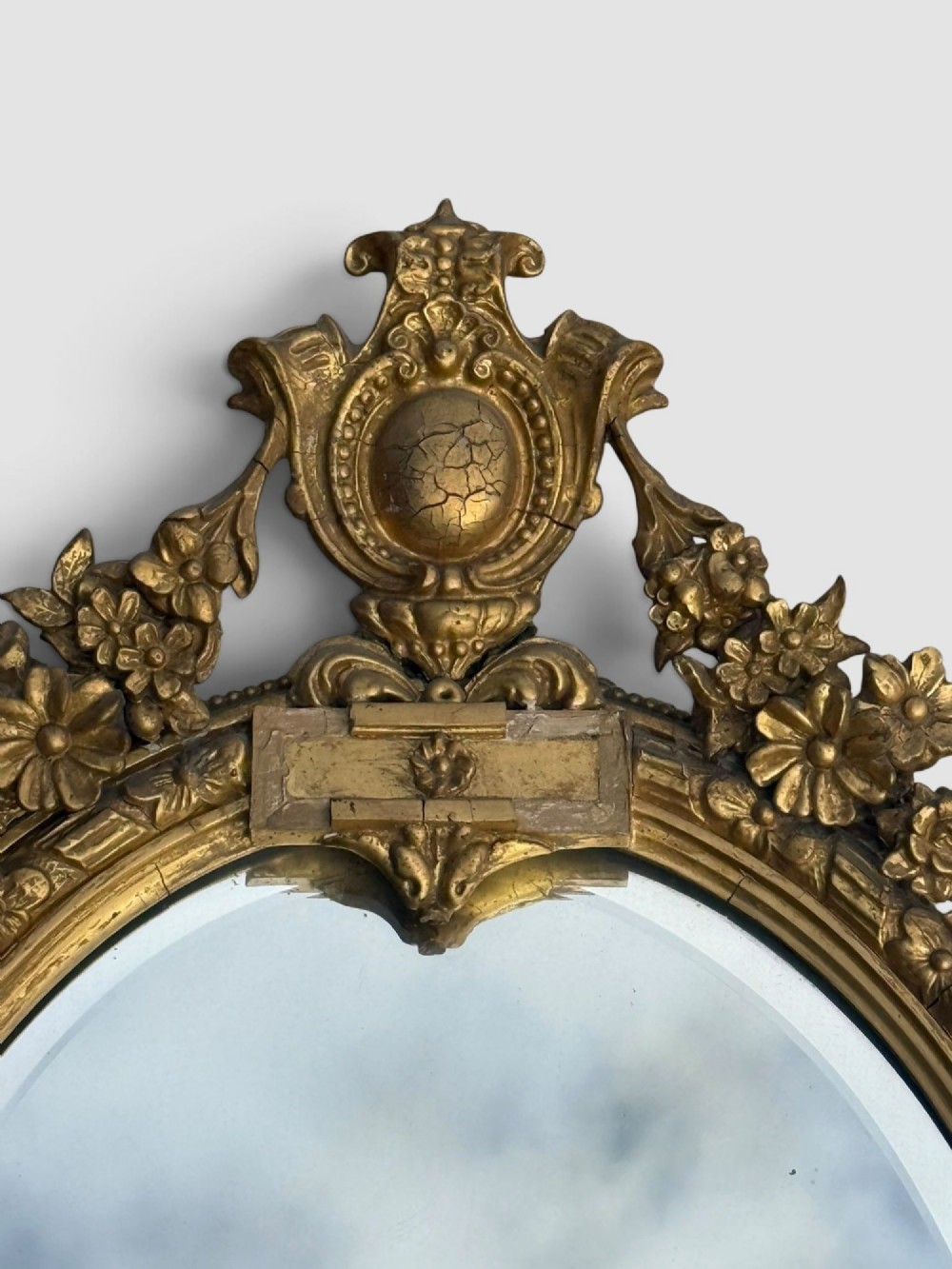 An oval Victorian gilt gesso wall mirror featuring an ornate floral crest, decorative scrollwork, and bevelled mirror glass. View VII
