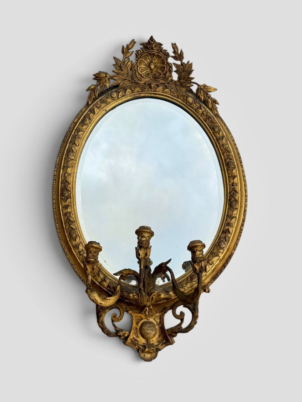 An oval Victorian giltwood and gesso girandole wall mirror featuring floral cresting, a beaded inner rim, and a three-branch candle sconce at the base.