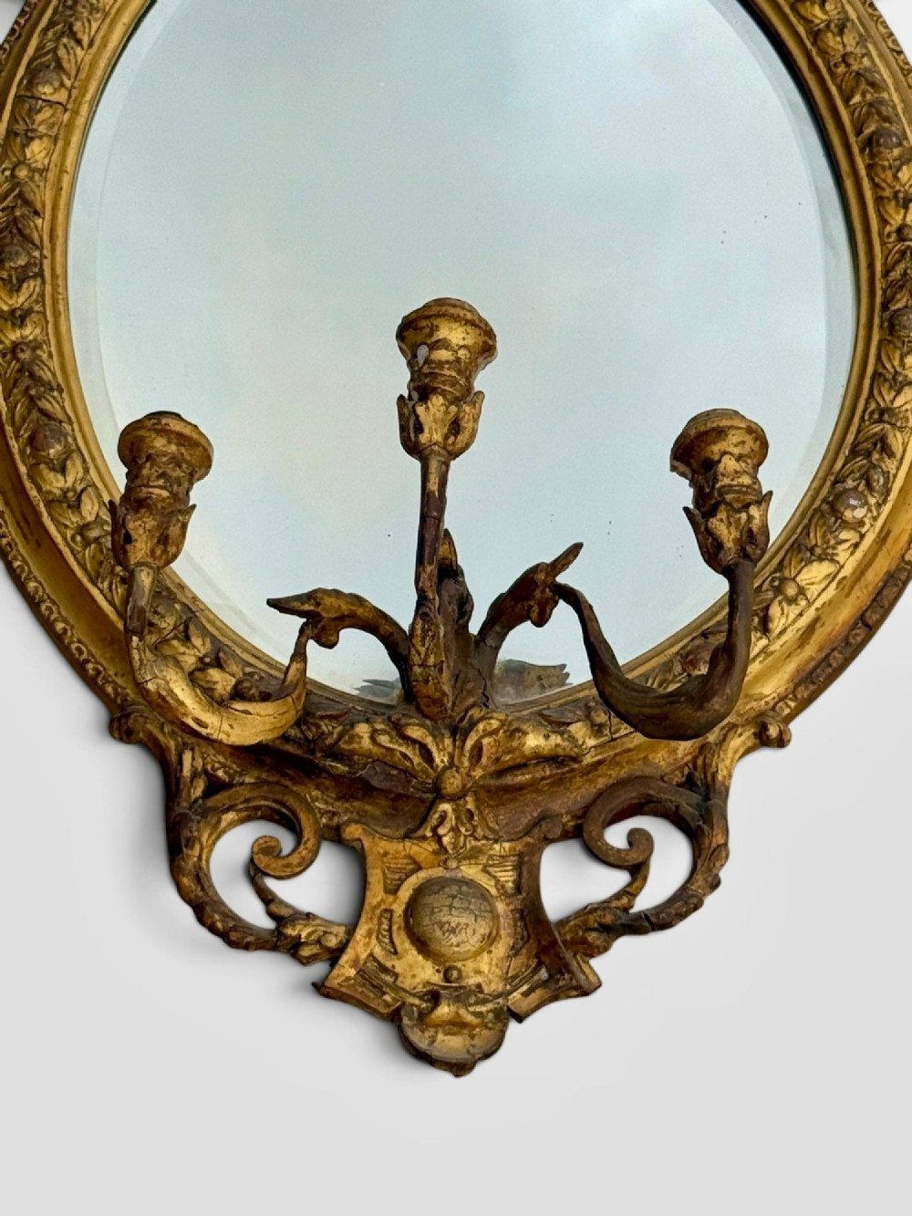 An oval Victorian giltwood and gesso girandole wall mirror featuring floral cresting, a beaded inner rim, and a three-branch candle sconce at the base. View IX