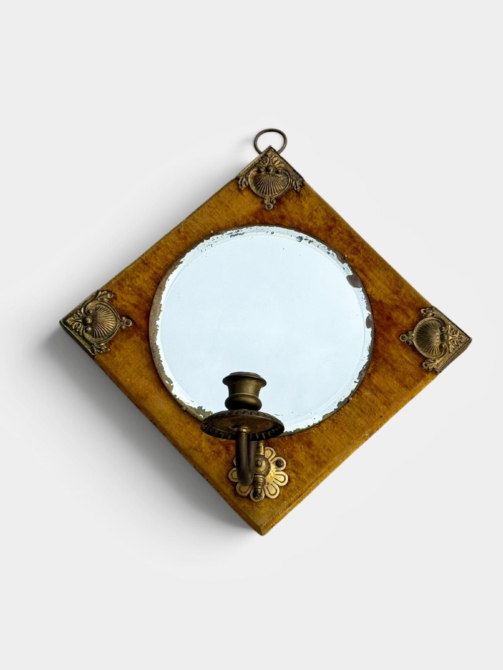 A diamond-shaped wall mirror featuring a mustard yellow velvet frame with decorative brass corner mounts, a central round bevelled glass mirror, and an integrated brass candle sconce at the lower vertex. View VI