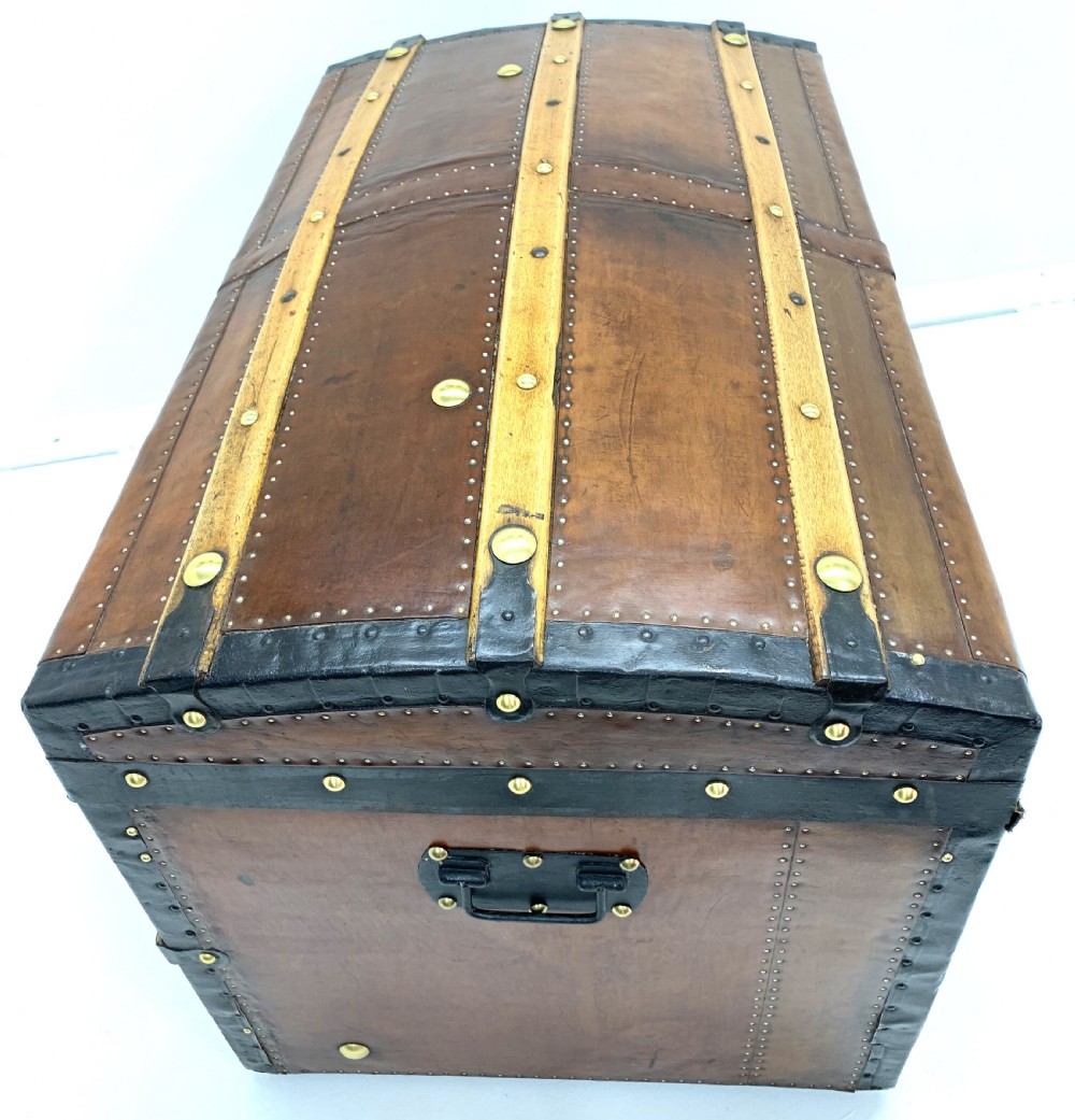 An antique leather-bound dome top steamer trunk with brass studs, wooden slats, and a central brass lock mechanism. View X