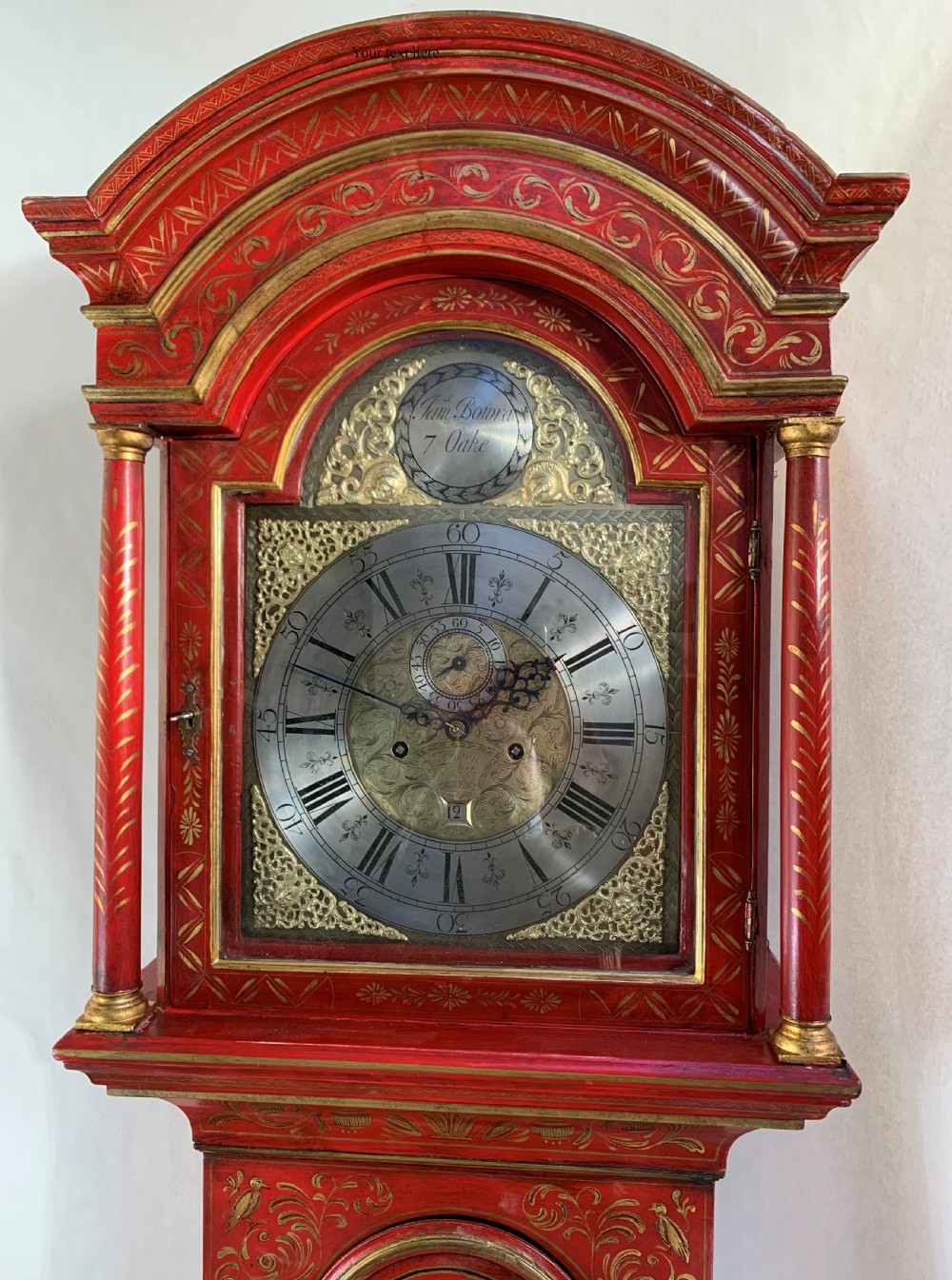 An antique red lacquered longcase clock decorated with gilded chinoiserie landscape scenes, featuring an arched hood and brass dial. View II