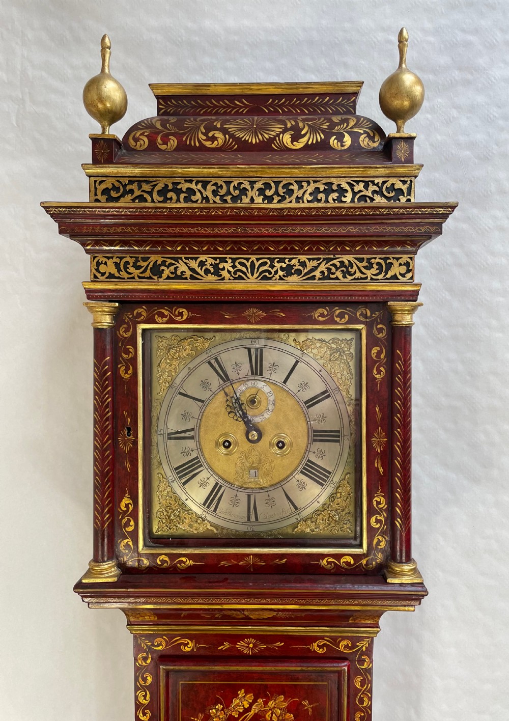 A red lacquered longcase clock featuring detailed gilt chinoiserie scenes, brass finials, and an engraved brass dial. View II