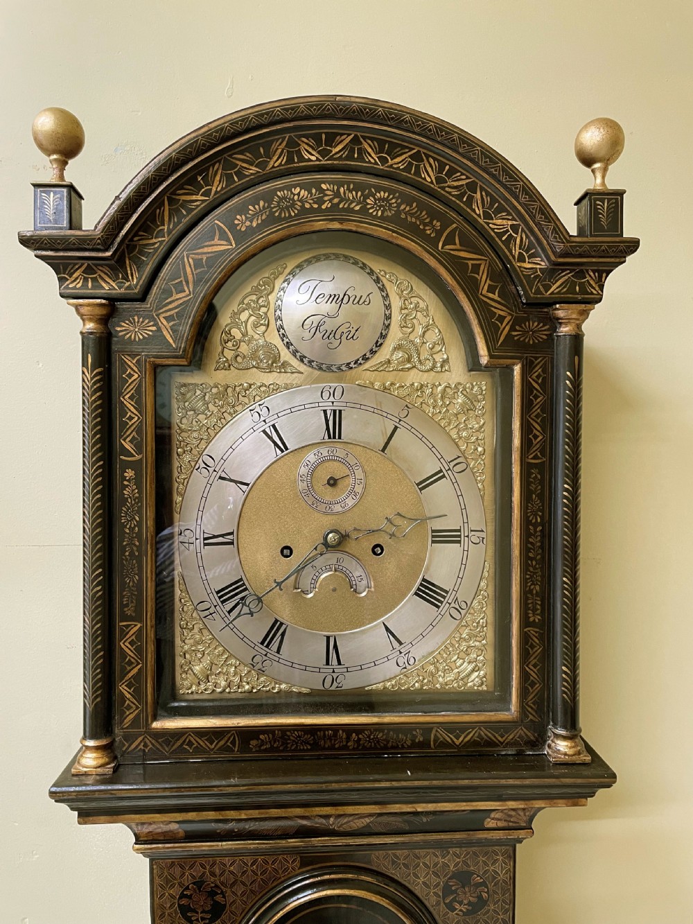 An antique dark green japanned longcase clock featuring gilded chinoiserie landscape details, an arched hood with gold finials, and a brass dial with a Tempus Fugit boss. View II