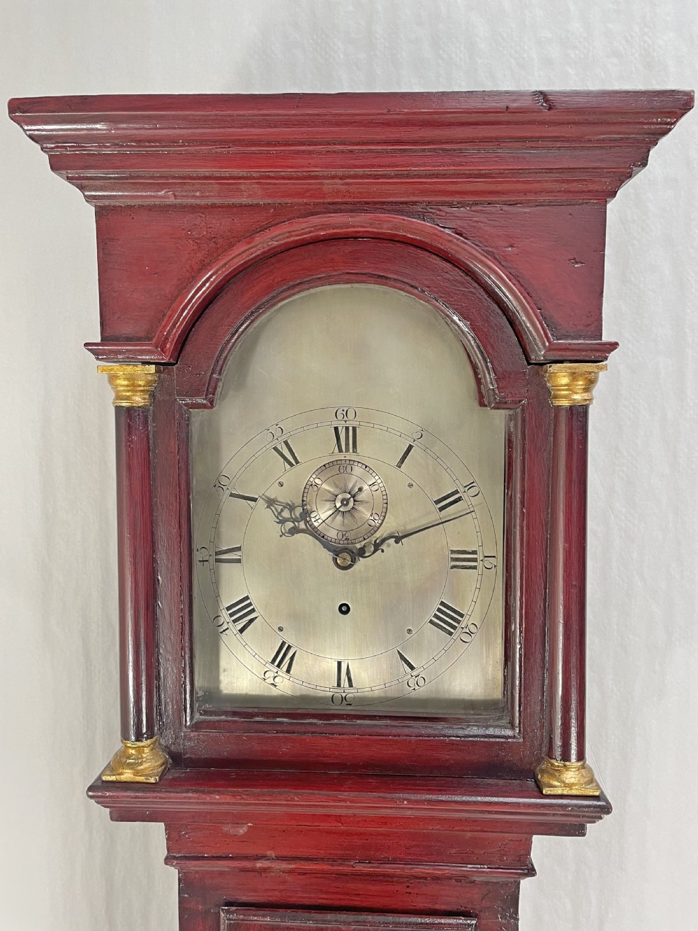 A tall antique longcase clock featuring a deep red painted wooden finish, an arched clock dial with Roman numerals, and gilded column capitals. View II