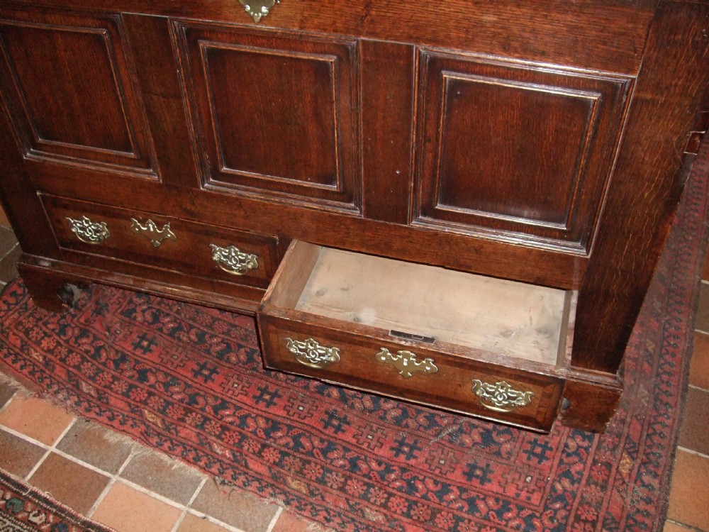 An 18th-century dark oak mule chest featuring a hinged top lid, three fielded panels across the front facade, two base drawers with brass handles, and bracket feet. View II