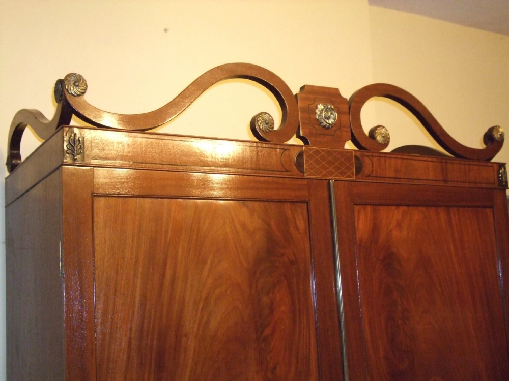An Irish Regency mahogany linen press featuring a decorative scrolled swan-neck pediment, twin cupboard doors with flame mahogany panels, and a chest of drawers below. View II