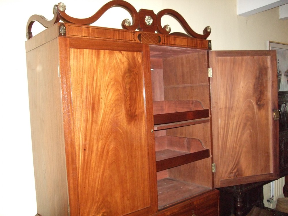 An Irish Regency mahogany linen press featuring a decorative scrolled swan-neck pediment, twin cupboard doors with flame mahogany panels, and a chest of drawers below. View III