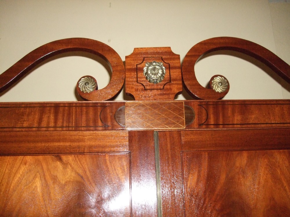 An Irish Regency mahogany linen press featuring a decorative scrolled swan-neck pediment, twin cupboard doors with flame mahogany panels, and a chest of drawers below. View IV
