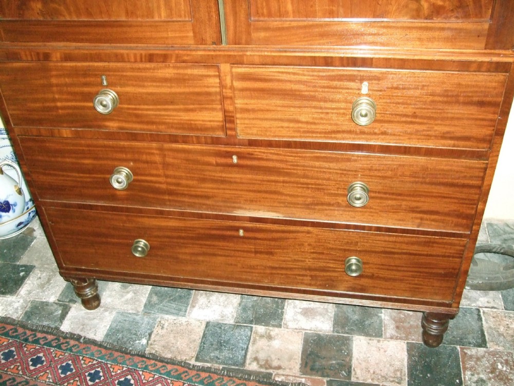 An Irish Regency mahogany linen press featuring a decorative scrolled swan-neck pediment, twin cupboard doors with flame mahogany panels, and a chest of drawers below. View V