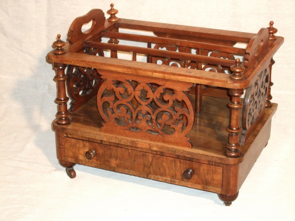 A Victorian walnut music Canterbury featuring open compartments with carved fretwork dividers, corner turned pillars with finials, a lower drawer with twin knobs, and castor feet.