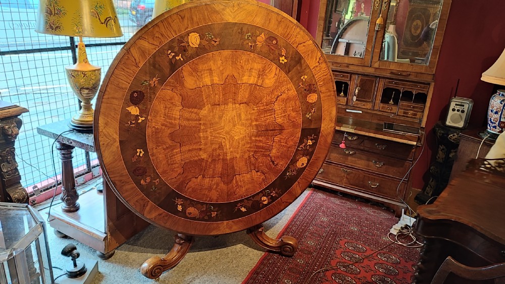 A circular Victorian burr walnut tilt-top centre table featuring floral marquetry inlay around the rim and carved scroll legs.