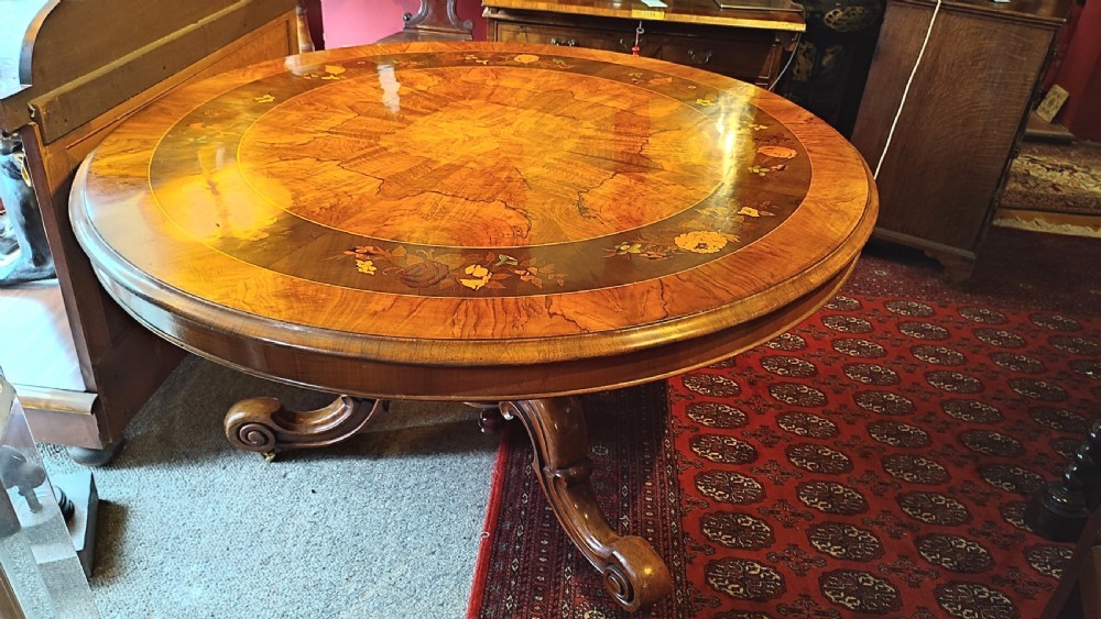 A circular Victorian burr walnut tilt-top centre table featuring floral marquetry inlay around the rim and carved scroll legs. View IV