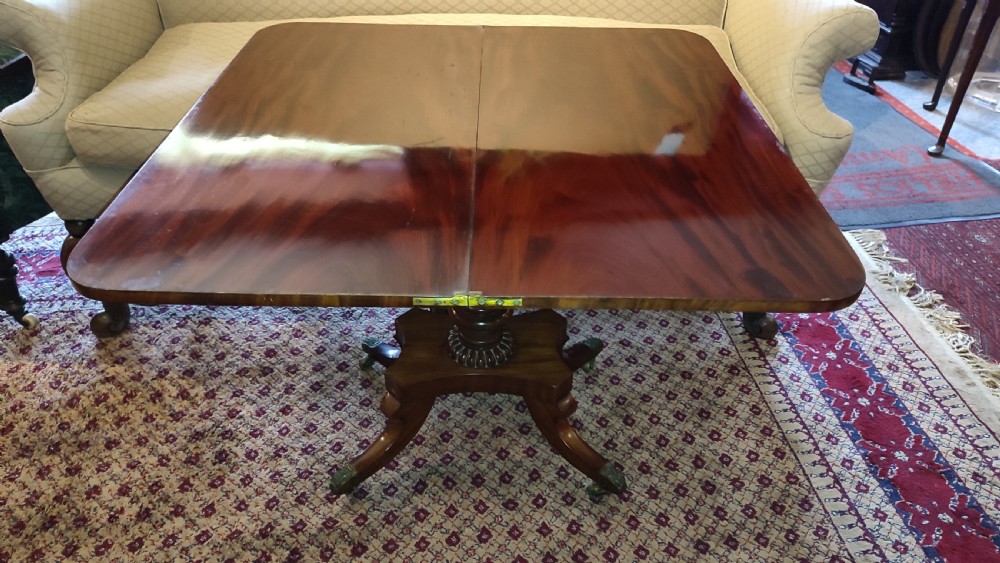 A Regency period mahogany tea table featuring a polished fold-over rectangular top set on a carved central pedestal and four splayed legs with brass castors. View IV