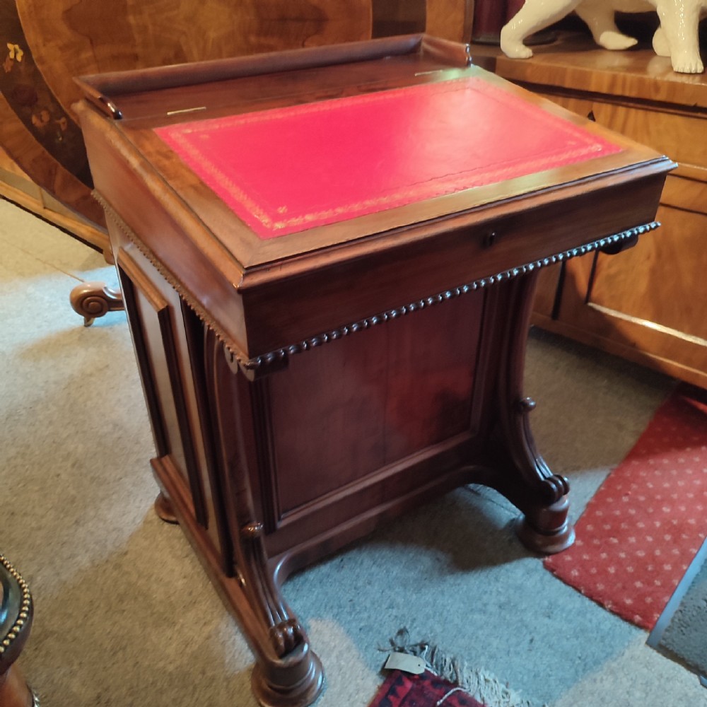 A Regency mahogany davenport desk featuring a red leather-lined sloped writing top, carved scroll supports, and beaded edging.