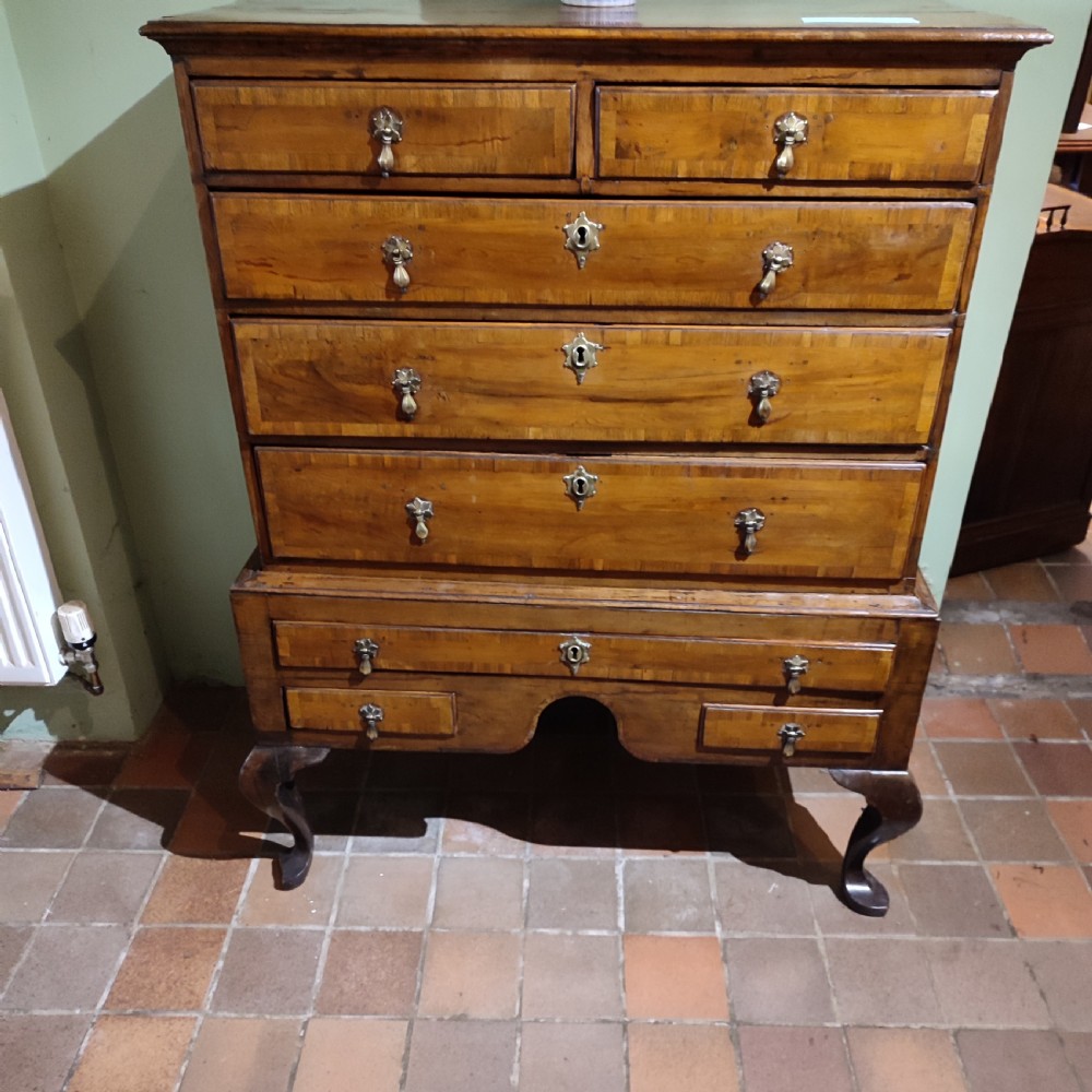 An antique Queen Anne yew wood chest on stand with crossbanded drawers, brass drop handles, and an arched apron raised on cabriole legs.