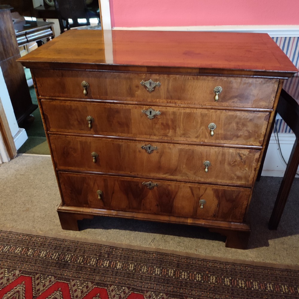 A George I period warm brown walnut chest of drawers featuring figured grain, crossbanded drawer fronts, brass drop handles, and bracket feet.