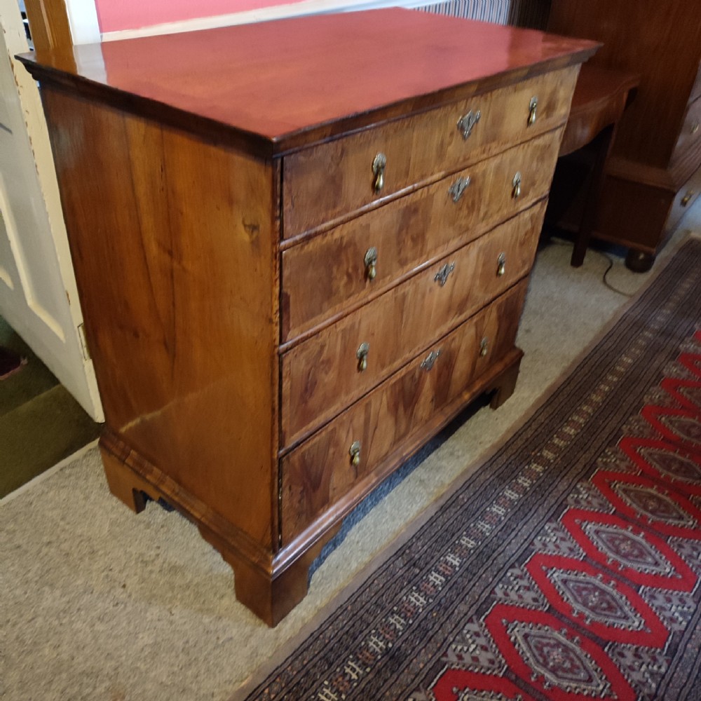 A George I period warm brown walnut chest of drawers featuring figured grain, crossbanded drawer fronts, brass drop handles, and bracket feet. View II