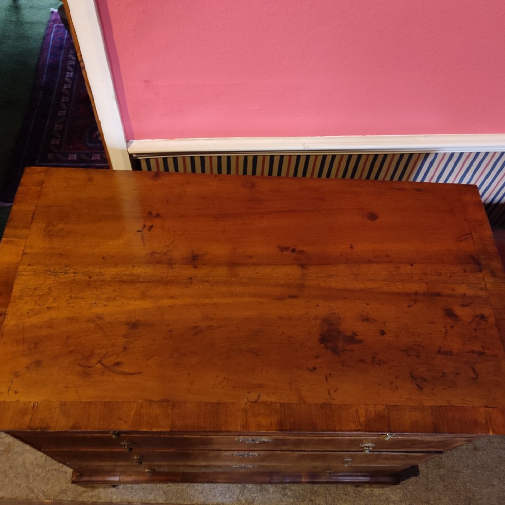 A George I period warm brown walnut chest of drawers featuring figured grain, crossbanded drawer fronts, brass drop handles, and bracket feet. View V