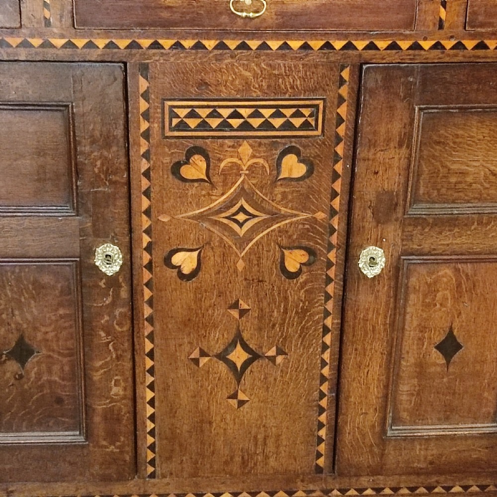A seventeenth-century inlaid oak court cupboard featuring geometric marquetry, turned supports, and the carved date 1678 on the upper frieze. View II