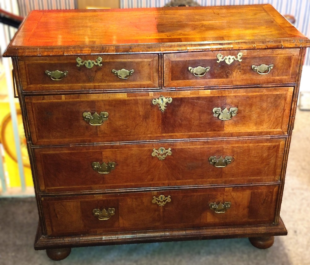 An early eighteenth-century walnut chest of drawers featuring decorative brass handles, crossbanded drawer fronts, and turned bun feet, constructed in two separate upper and lower sections.